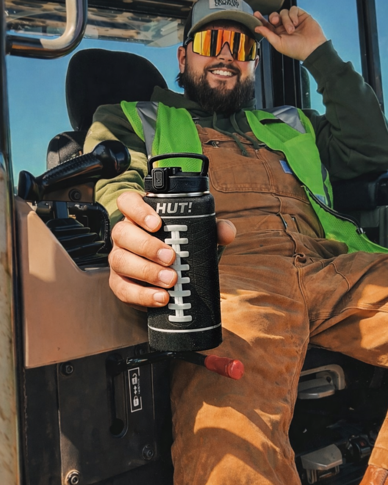 Person holding a black water bottle with a green shirt and brown overalls inside a vehicle.