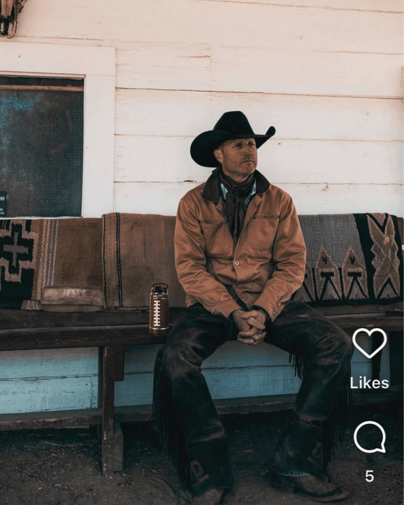 Man in a cowboy hat sitting on a bench with a water bottle beside him, in front of a wooden wall.