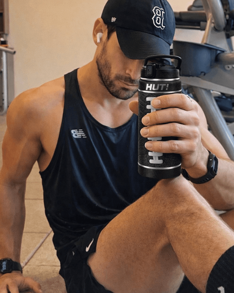 Man in gym wearing a black tank top and cap, holding a black 'HUT!' shaker bottle.