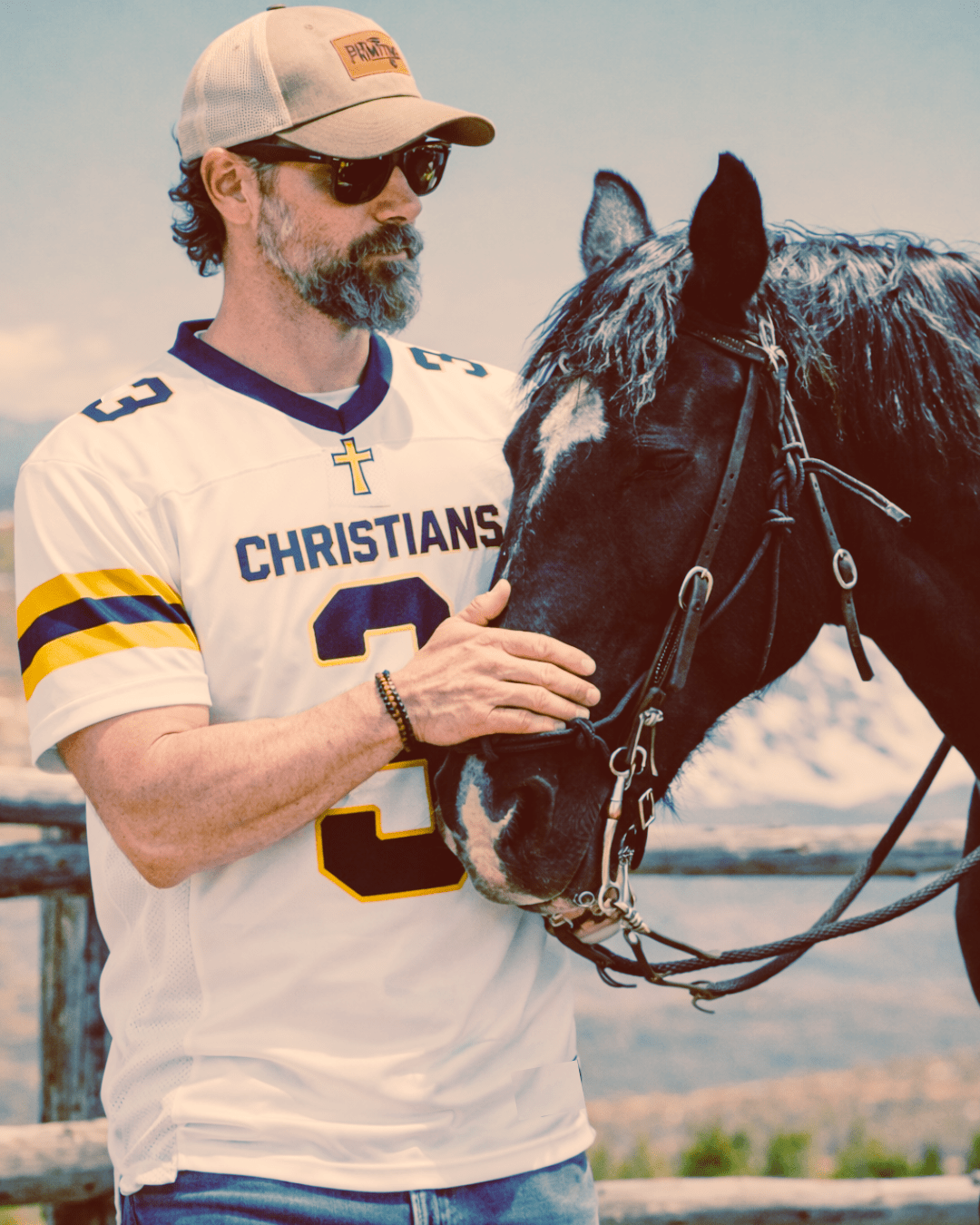 Man wearing a sports jersey and cap, holding a horse's bridle with a scenic background